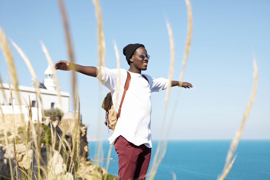 Young black man wearing trendy hipster clothes standing on rocks overlooking sea, spreading his arms, feeling carefree and happy, smiling, breathing fresh air. People, lifestyle and traveling